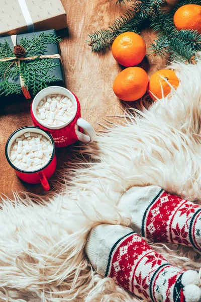 Vista parcial de la mujer en calcetines sobre fondo con tazas de bebida de cacao, mandarinas, galletas y regalos de Navidad - foto de stock