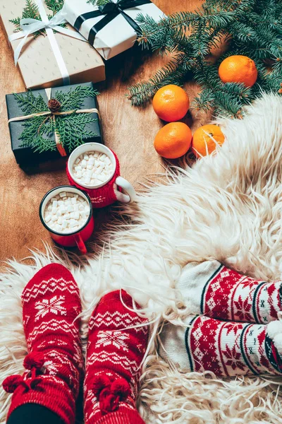 Cropped shot of women in winter socks at background with wrapped christmas presents and cups of hot chocolate with marshmallows — Stock Photo