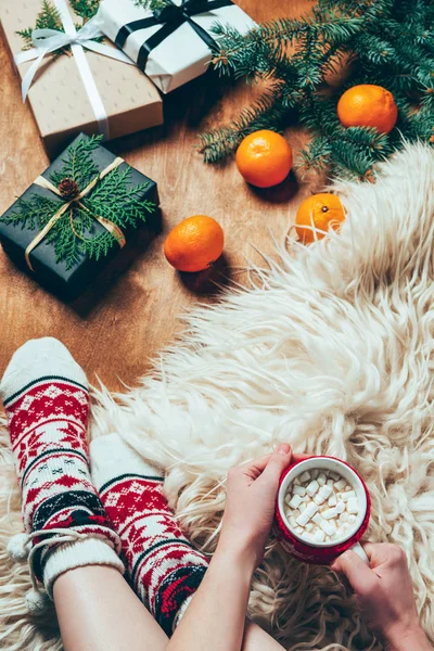 Частковий вид жінки з чашкою гарячого шоколаду та зефіру на фоні з різдвяними подарунками та мандаринами — стокове фото