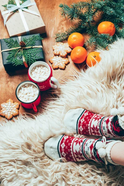 Vista parcial de la mujer en calcetines sobre fondo con tazas de bebida de cacao, mandarinas, galletas y regalos de Navidad - foto de stock