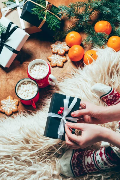 Teilansicht einer Frau mit Weihnachtsgeschenk auf Hintergrund mit Tassen mit Kakaogetränken, Keksen und Mandarinen — Stockfoto