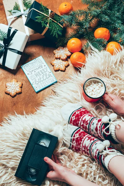 Teilansicht einer Frau mit Tasse Heißgetränk und Videokassette auf Hintergrund mit Plätzchen, Mandarinen und Weihnachtsgeschenken — Stockfoto