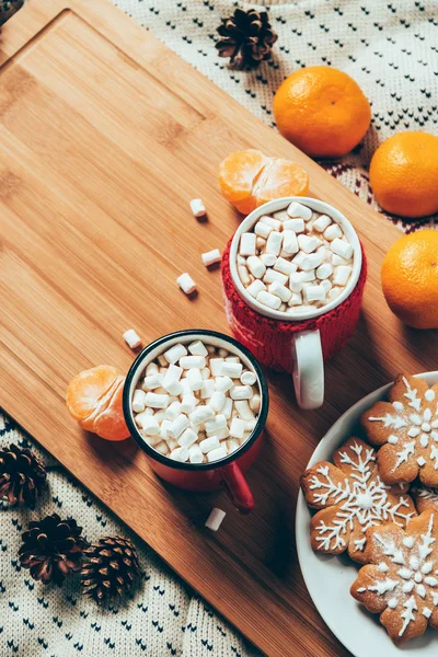 Draufsicht auf Tassen mit heißer Schokolade mit Marshmallows, Plätzchen und Mandarinen auf Deckenhintergrund, weihnachtliches Frühstückskonzept — Stockfoto
