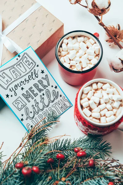 Vista superior de la postal con tener que casarse con pequeñas letras de Navidad, presente y tazas de bebidas de cacao con malvaviscos en la mesa blanca - foto de stock