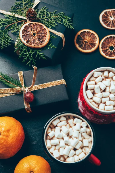 Top view of cups of hot drinks with marshmallows, tangerines and christmas presents on black surface — Stock Photo