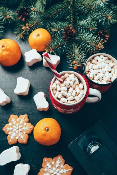 Vista superior de copos de bebidas quentes com marshmallows, cassete de vídeo e tangerinas na superfície preta, conceito de café da manhã de natal — Fotografia de Stock