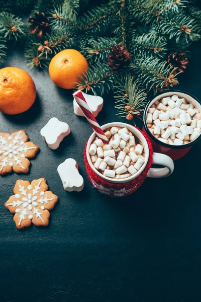 Vista superior de las tazas de bebidas calientes con malvaviscos, galletas y mandarinas en la superficie negra, concepto de desayuno de Navidad — Stock Photo