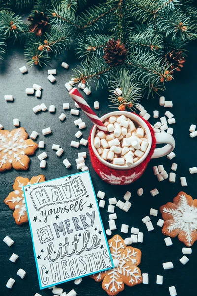 Piatta giaceva con tazza di bevanda calda con marshmallow, biscotti e cartolina con te allegro piccolo Natale lettering su tavolo nero — Foto stock