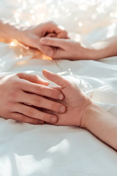 Vista parziale di coppia che si tiene per mano mentre si trova a letto con ghirlanda — Foto stock