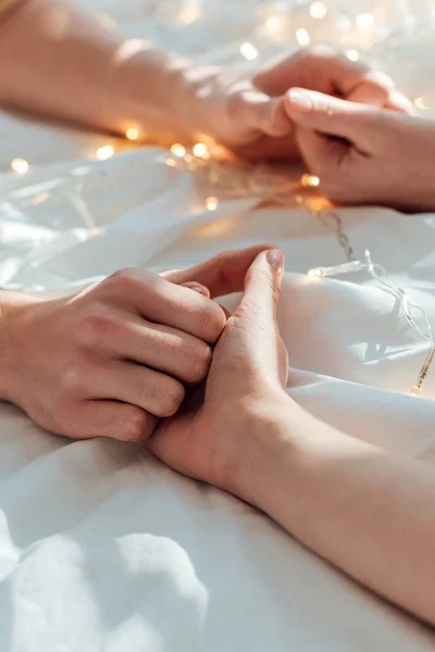 Vue partielle du couple tenant la main tout en étant couché au lit avec une guirlande — Photo de stock