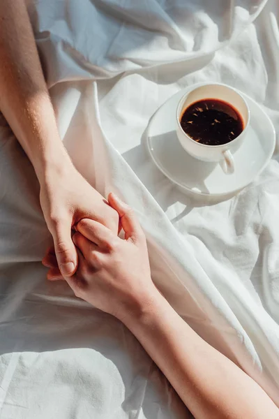 Recortado disparo de pareja cogida de la mano en la sábana blanca con taza de café aromático - foto de stock