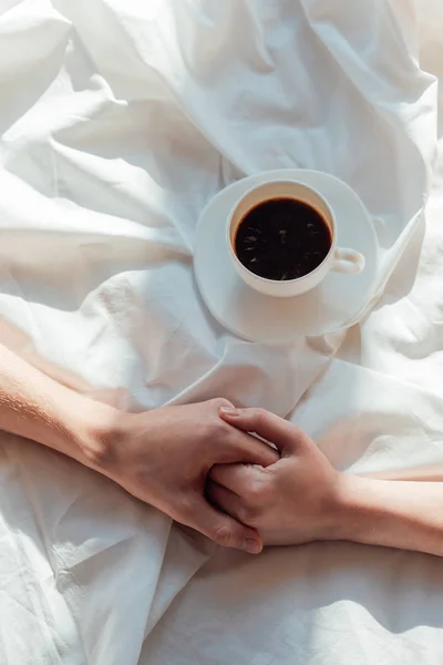 Cropped shot of couple holding hands on white bed sheet with cup of aromatic coffee — Stock Photo