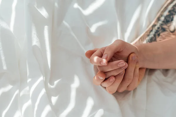 Частковий вид люблячої пари, що тримає руки, лежачи на білому простирадлі — стокове фото