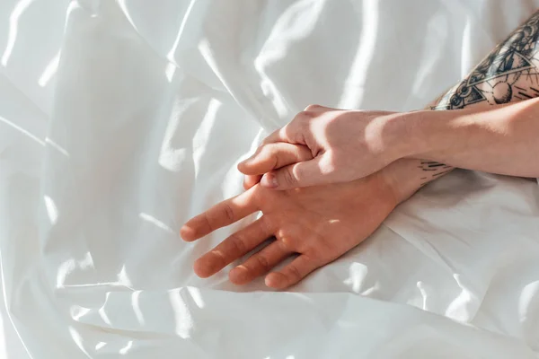 Visão parcial do casal amoroso de mãos dadas enquanto deitado em lençol branco — Fotografia de Stock