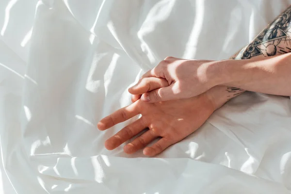 Partial view of loving couple holding hands while lying on white bed sheet — Stock Photo