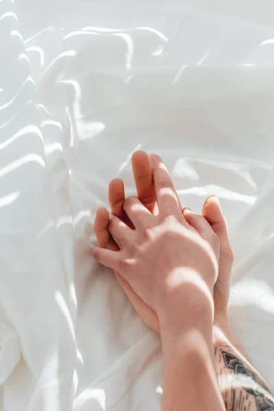 Partial view of loving couple holding hands while lying on white bed sheet — Stock Photo