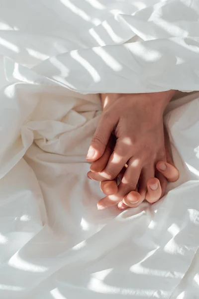 Vista parcial de pareja amorosa cogida de la mano mientras yacen bajo una manta juntos - foto de stock