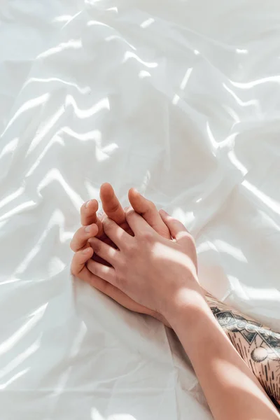 Частичный вид любящей пары, держащейся за руки, лежащей на белом простыне — стоковое фото