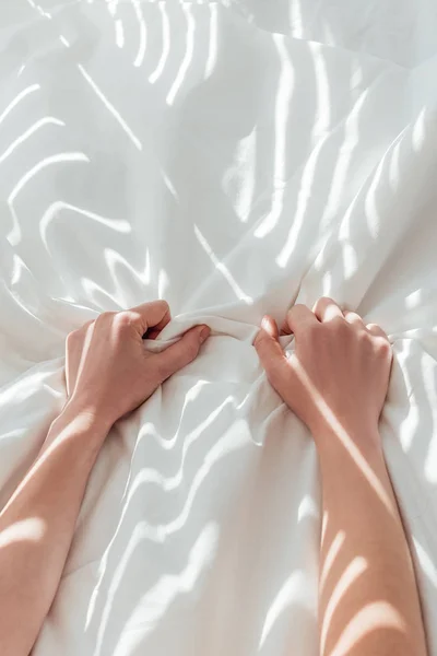 Vista parcial de la mujer sosteniendo sábana blanca mientras está acostada en la cama - foto de stock