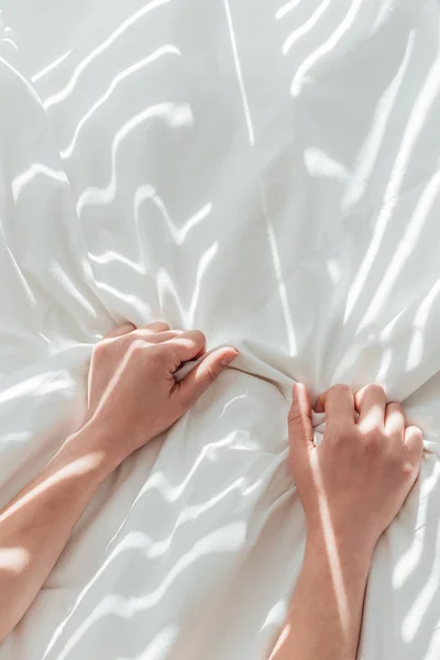 Vista parcial de la mujer sosteniendo sábana blanca mientras está acostada en la cama - foto de stock