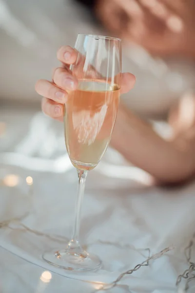 Vue partielle de la femme tenant un verre de champagne sur drap de lit blanc avec guirlande — Photo de stock