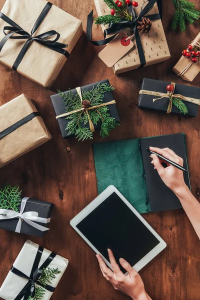 Частковий вид жінки з використанням планшета і списку письма в щоденнику на дерев'яному столі з різдвяними подарунками — стокове фото
