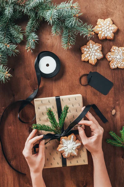 Geschnittene Ansicht einer Frau, die Weihnachtsgeschenk mit Schleife und Tannenzweigen auf Holzgrund mit Schneeflockenplätzchen dekoriert — Stockfoto