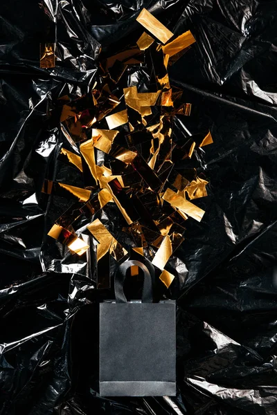 Vista superior de la bolsa de compras de papel con confeti dorado sobre fondo de papel de embalaje negro, concepto de viernes negro - foto de stock