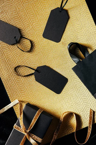 Plano con etiquetas de precio en blanco, bolsa de compras y regalo con cinta dorada sobre fondo negro y dorado, concepto de viernes negro — Stock Photo