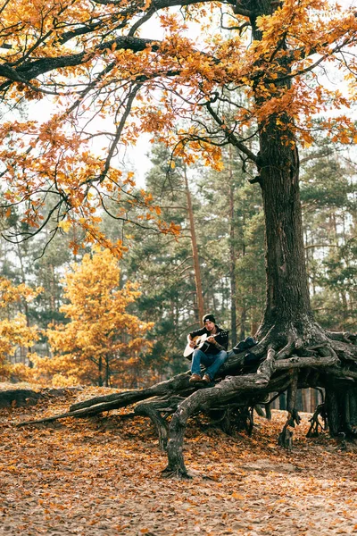 Adult male playing acoustic guitar sitting on tree in park — Stock Photo