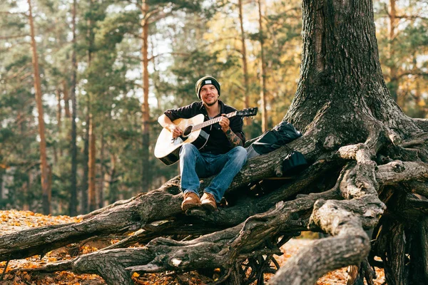 Adulte mâle jouant guitare acoustique assis sur l'arbre dans le parc — Photo de stock
