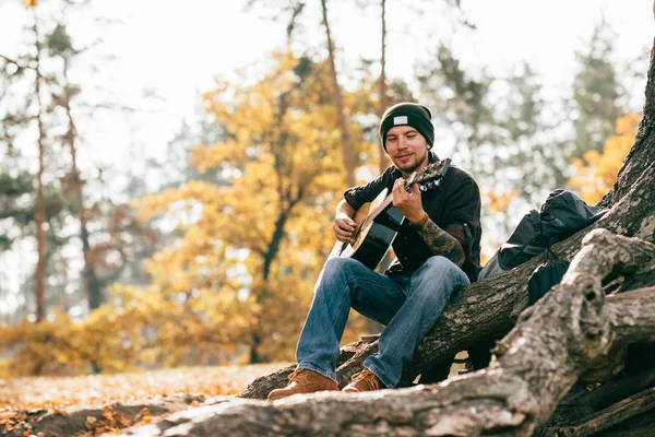 Adulte mâle jouant guitare acoustique assis sur l'arbre dans le parc — Photo de stock