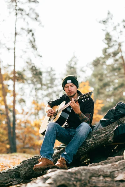 Adulte mâle jouant guitare acoustique assis sur l'arbre dans le parc — Photo de stock