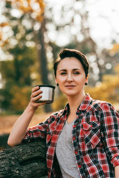 Доросла жінка дивиться на камеру ходіння металевої чашки термос на розмитому автентичному фоні — стокове фото