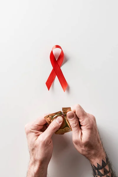 Male hands opening golden packaging of condom with aids awareness red ribbon on white background — Stock Photo