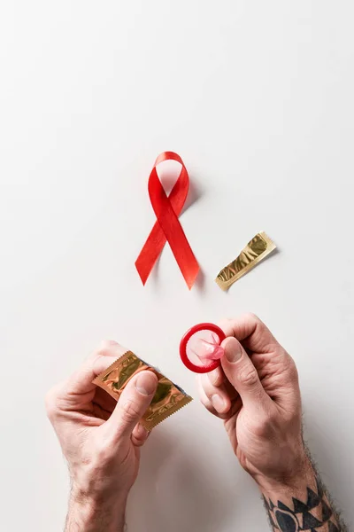Manos masculinas que sostienen el embalaje dorado y el condón rojo con la cinta roja del conocimiento de las ayudas en el fondo blanco - foto de stock