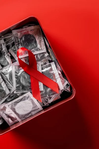 Vue du dessus du ruban rouge de sensibilisation aux aides et des préservatifs en argent en boîte sur fond rouge — Photo de stock
