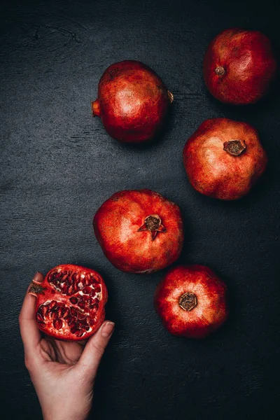 Partial view of woman holding cut pomegranate on black surface with arranged wholesome garnets — Stock Photo