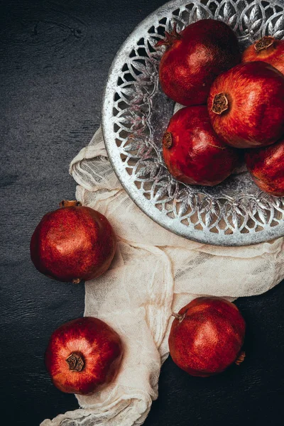 Food composition with ripe and fresh pomegranates, metal bowl and gauze on black tabletop — Stock Photo
