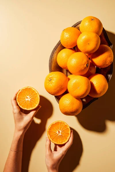 Foto recortada de mujer sosteniendo mitades de mandarina sobre fondo beige con cuenco de metal - foto de stock