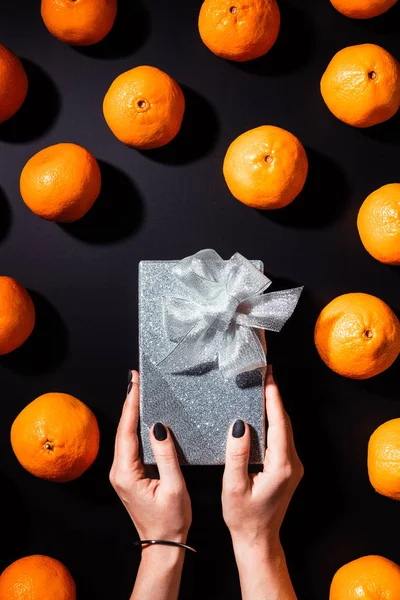 Vista parcial da mulher segurando caixa de presente de prata na mesa preta com tangerinas frescas ao redor — Fotografia de Stock