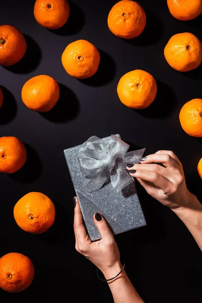 Vista parcial da mulher segurando caixa de presente de prata na mesa preta com tangerinas frescas ao redor — Fotografia de Stock