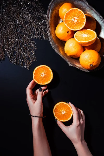 Visão parcial da mulher segurando metades de tangerina na superfície preta com galho de prata decorativo e tangerinas em tigela de metal — Fotografia de Stock