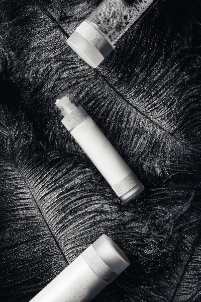 Deitado plano com loção, creme de beleza e água micelar na superfície com penas pretas cobertas de prata — Fotografia de Stock