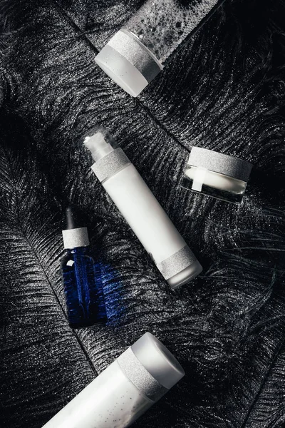 Vue surélevée de crème de beauté, lotion, sérum et eau micellaire à la surface avec des plumes noires recouvertes d'argent — Photo de stock