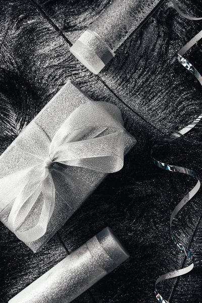 Top view of christmas gift box, lotion, micellar water and ribbon on surface with black feathers covered by silver — Stock Photo