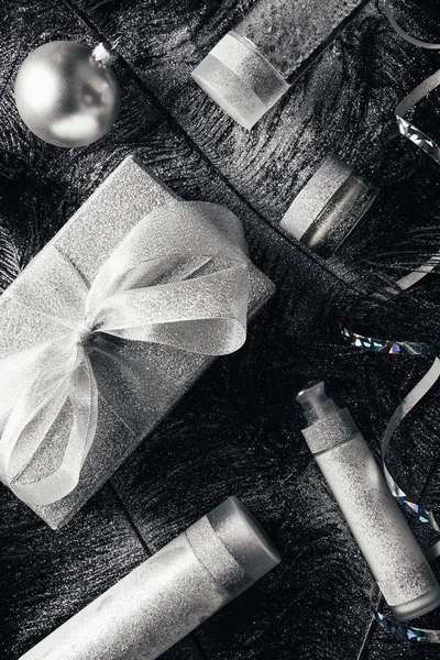 Vista elevada de la bola de Navidad, caja de regalo y equipo de tratamiento de belleza en la superficie con plumas negras cubiertas de plata - foto de stock