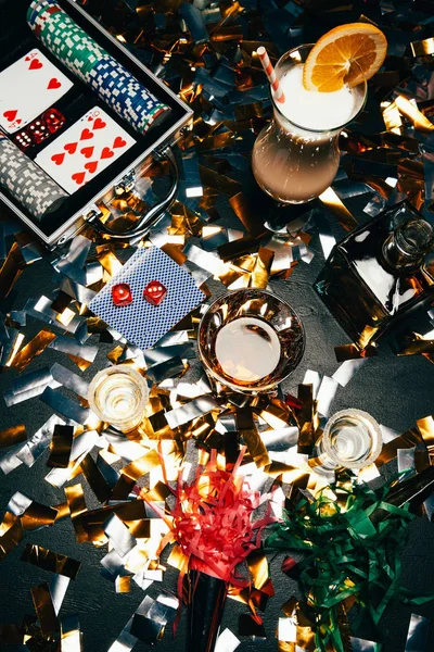 Vista elevada de coquetéis alcoólicos, cartas de jogo, fichas de poker e chifres de festa na mesa coberta por confetes dourados — Fotografia de Stock