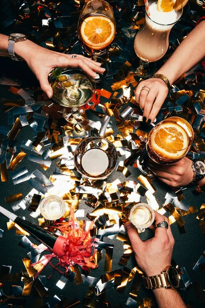 Cropped image of friends with luxury watches celebrating alcohol at table covered by golden confetti — Stock Photo