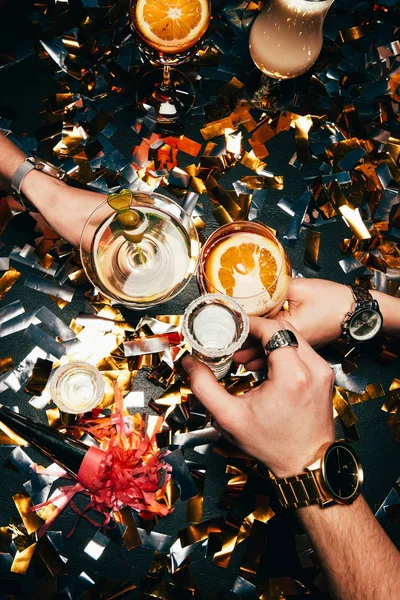 Vista parcial de amigos con relojes de lujo tintineo por vasos con alcohol sobre la mesa cubierta por confeti de oro - foto de stock